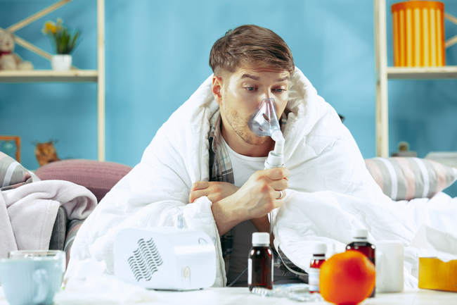 523 bearded sick man with flue sitting on sofa at home covered with warm blanket and using an inhaler when coughing